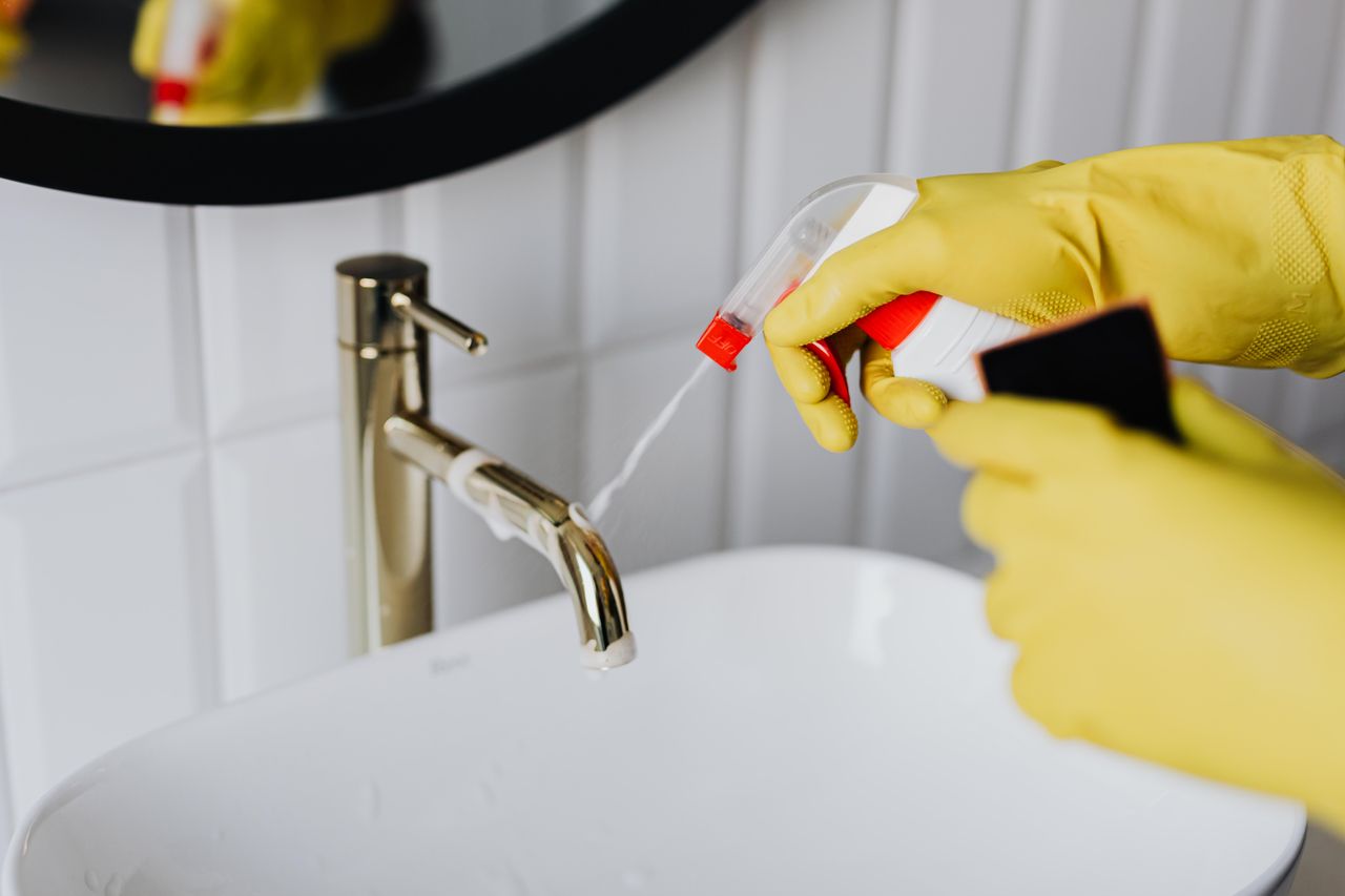 From above, an anonymous housekeeper in yellow rubber gloves sprays cleaner onto a chrome bathroom water faucet